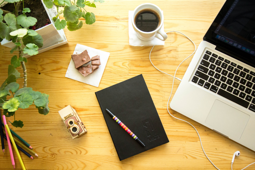 This image shows a flatlay of a notebook, a laptop, coffee, and other items against a light wood table.